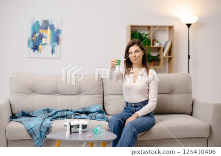 Young woman wearing casual clothing, using an analgesic spray to soften the throat, sitting on a sofa in a home interior Young woman wearing casual clothing, using an analgesic spray to soften the throat, sitting on a sofa in a home interior 125410406