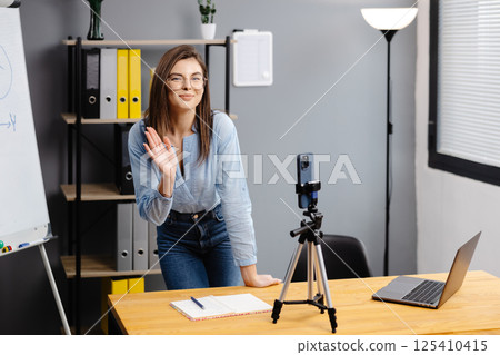 Positive blogger woman speaking at smartphone webcam on tripod, recording post for vlog. Online teacher, coach working, giving webinar at home. Presenter taking video on cellphone 125410415