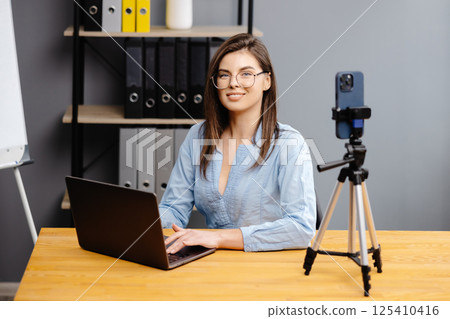 Videoconference And Training. Class of diverse students learning remotely online using web app. Female teacher providing lesson with laptop, recording video on cellphone on tripod Videoconference And Training. Class of diverse students learning remotely online using web app. Female teacher providing lesson with laptop, recording video on cellphone on tripod 125410416