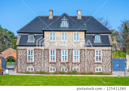 A historic building stands amidst vibrant greenery in Copenhagen's Rosenborg Castle Garden. The sun lights up its charming facade, showcasing traditional architectural details and tranquility. 125411260