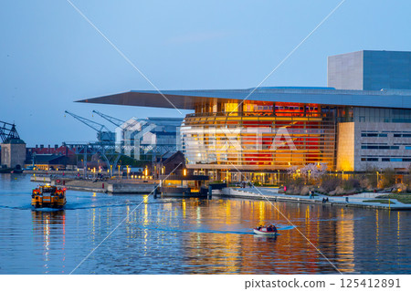 Visitors enjoy a serene evening at Copenhagen Opera House as boats navigate the calm waters of the harbor. The building's lights reflect beautifully on the water's surface. 125412891