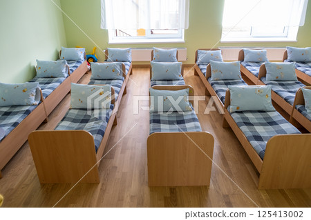 Many small beds with fresh linen in daycare preschool empty bedroom interior for comfortable afternoon nap of the kids. Many small beds with fresh linen in daycare preschool empty bedroom interior for comfortable afternoon nap of the kids. 125413002