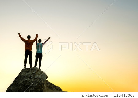 Man and woman hikers standing on a big stone at sunset in mountains. Couple raising up hands on high rock in evening nature. Tourism, traveling and healthy lifestyle concept. Man and woman hikers standing on a big stone at sunset in mountains. Couple raising up hands on high rock in evening nature. Tourism, traveling and healthy lifestyle concept. 125413003