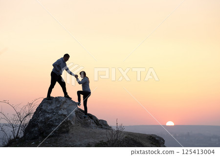 Man and woman hikers helping each other to climb stone at sunset in mountains. Couple climbing on high rock in evening nature. Tourism, traveling and healthy lifestyle concept. Man and woman hikers helping each other to climb stone at sunset in mountains. Couple climbing on high rock in evening nature. Tourism, traveling and healthy lifestyle concept. 125413004