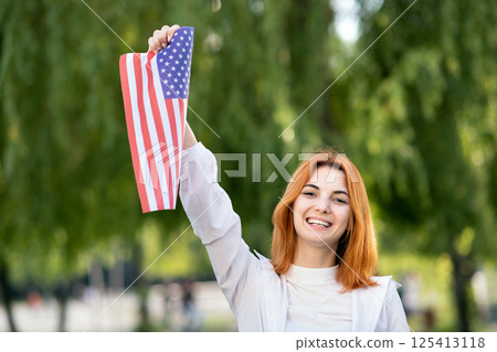 Happy young woman posing with USA national flag holding it in her outstretched hand standing outdoors in summer park. Pretty girl celebrating United States independence day. Happy young woman posing with USA national flag holding it in her outstretched hand standing outdoors in summer park. Pretty girl celebrating United States independence day. 125413118