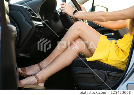 Close up of young woman driver legs in yellow summer dress behind steering wheel driving a car. Close up of young woman driver legs in yellow summer dress behind steering wheel driving a car. 125413608