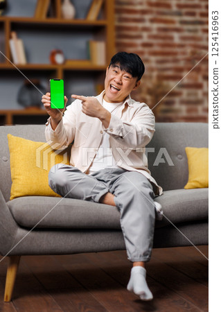 Amazing Useful App, Great Mobile Offer, Mobile Advertisement, Excited man seated on a gray couch happily points to a smartphone displaying a green screen. The cozy living room features yellow cushions 125416963