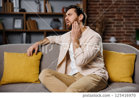 severe toothache, Upset man, holds his cheek, man is sitting on a gray couch in a warm-toned room, visibly troubled by jaw pain. He holds his face with one hand, suggesting distress, while surrounded severe toothache, Upset man, holds his cheek, man is sitting on a gray couch in a warm-toned room, visibly troubled by jaw pain. He holds his face with one hand, suggesting distress, while surrounded 125416970