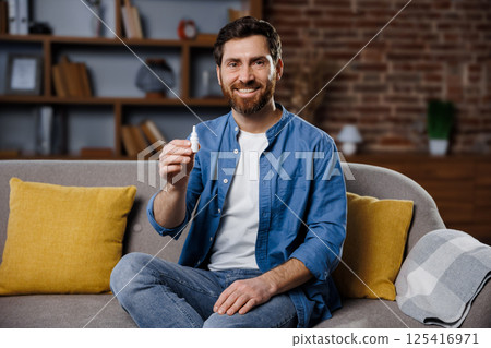 eye drops, portrait smiling man, health care, smiling man sits comfortably on a couch in a modern living room, showcasing a small bottle. The room features rustic decor, shelves, and warm colors eye drops, portrait smiling man, health care, smiling man sits comfortably on a couch in a modern living room, showcasing a small bottle. The room features rustic decor, shelves, and warm colors 125416971