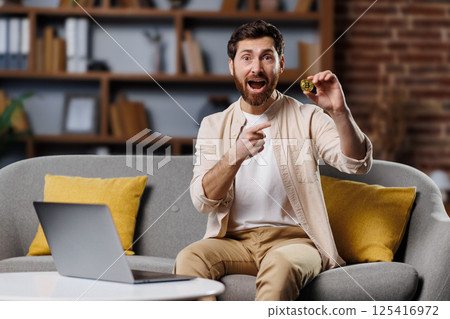 excited man, shows cryptocurrency, gold bitcoin coin, man excitedly holds up a freshly baked cookie while seated on a soft couch, surrounded by colorful cushions. excited man, shows cryptocurrency, gold bitcoin coin, man excitedly holds up a freshly baked cookie while seated on a soft couch, surrounded by colorful cushions. 125416972