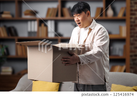 opening delivery box, excited man, Online shopping concept, man looks excited as he opens a large cardboard box in a comfortable living room. The surrounding shelves are filled with books, creating an 125416974