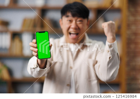 Excited surprised man, winning lottery, phone screen, surprised, asian man, joyful person holds up a smartphone with a vibrant green screen, expressing happiness and triumph in a cozy environment Excited surprised man, winning lottery, phone screen, surprised, asian man, joyful person holds up a smartphone with a vibrant green screen, expressing happiness and triumph in a cozy environment 125416984