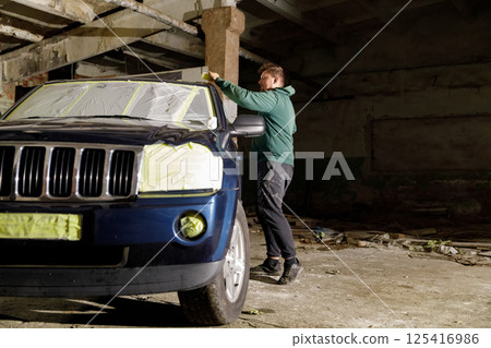 A worker is preparing a Jeep for refinishing inside an abandoned warehouse. The vehicle is covered in masking tape, and debris is scattered around the dimly lit space. 125416986