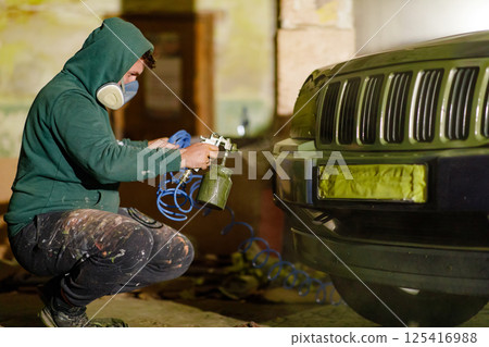 A person is focused on painting a vehicle using a spray gun inside a dimly lit workshop. They are wearing a hoodie and mask, clearly working on refinishing the car's front end. 125416988