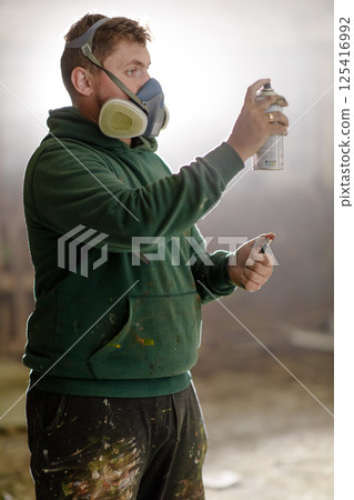 An artist uses spray paint to create a colorful piece in a workshop filled with art supplies. Dressed in a green hoodie and mask, he focuses on his craft in a misty setting. 125416992