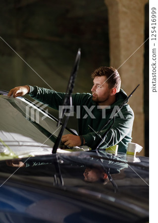 A man focuses on replacing the windshield of a car in a dimly lit area during nighttime. He is carefully adjusting the glass and ensuring a proper fit. A man focuses on replacing the windshield of a car in a dimly lit area during nighttime. He is carefully adjusting the glass and ensuring a proper fit. 125416999