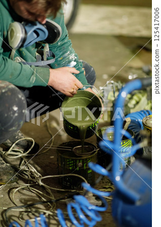 A focused worker in a paint-stained sweater carefully pours green paint from one container to another in a dimly lit workshop, surrounded by various tools and equipment. 125417006