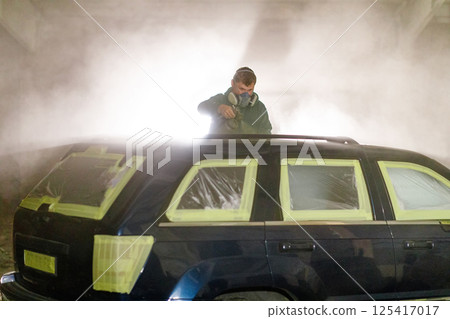 A man is applying paint to a black SUV in a workshop, using spray equipment while the car is masked with tape and plastic. The environment is misty, enhancing the focus on his work. 125417017