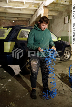In a poorly illuminated garage, a worker untangles blue coiled wires while surrounded by restoration tools and a vehicle ready for work. The atmosphere is focused and industrious. In a poorly illuminated garage, a worker untangles blue coiled wires while surrounded by restoration tools and a vehicle ready for work. The atmosphere is focused and industrious. 125417022