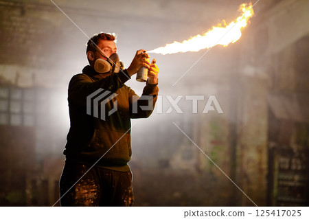 A man wearing a mask sprays paint that produces flames in a dimly lit, deserted warehouse. The atmosphere is intense as he showcases his creativity in this unique art form. A man wearing a mask sprays paint that produces flames in a dimly lit, deserted warehouse. The atmosphere is intense as he showcases his creativity in this unique art form. 125417025