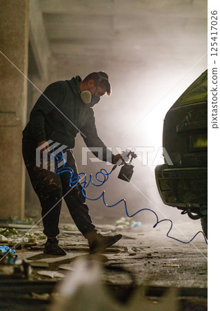 An artist works diligently in a neglected warehouse, applying colorful spray paint to a car. Dust fills the air as creativity flows in this urban art setting. 125417026