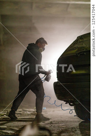 In a dimly lit workshop, a man wearing a mask is intently spray painting the rear of a car. Dust and mist create a moody atmosphere, highlighting the restoration work being performed. In a dimly lit workshop, a man wearing a mask is intently spray painting the rear of a car. Dust and mist create a moody atmosphere, highlighting the restoration work being performed. 125417044
