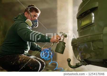 A person is carefully applying paint to a car using a spray gun while wearing a mask for protection. The workshop is dimly lit, highlighting the meticulous details of the process. 125417046