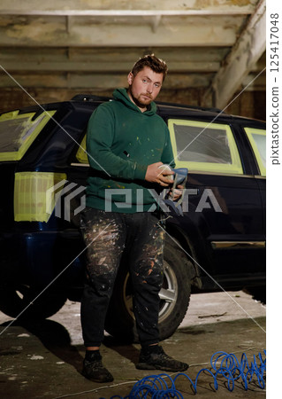 A young man stands beside a car in a garage, holding a spray gun. He wears a green hoodie and covered pants, indicating he has been working on the vehicle's restoration. 125417048