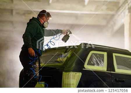 A skilled painter is carefully applying spray paint to a vehicle in a workshop. Wearing a mask for safety, he focuses on achieving a smooth finish while surrounded by protective materials. 125417056