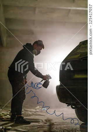 An artist applies spray paint to the back of a vintage car inside a dimly lit, abandoned warehouse, highlighting the blend of artistry and urban culture during evening hours. 125417057