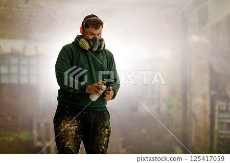 A focused artist prepares to spray paint in a deserted industrial location, surrounded by white mist and dim light, embodying creativity and urban artistry in a unique setting. 125417059