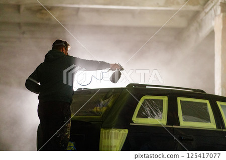 A person is spray painting a car in a dimly lit industrial workshop at night. The misty air and protective coverings create an artistic atmosphere while the vehicle is being prepped for a fresh coat. 125417077