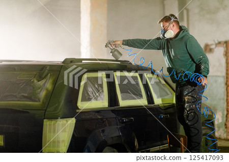 A worker in a mask sprays paint on a car's roof in a workshop. 125417093