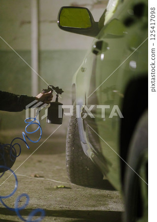 A technician is focused on refinishing a green car in a garage, utilizing a spray gun for precise touch-ups. The atmosphere is tinted with a soft glow, highlighting the meticulous work. 125417098
