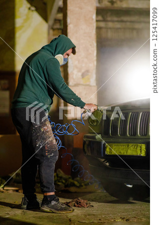 A worker in a green hoodie and mask is repairing a car using an air tool in a dimly lit, abandoned structure. Dust and debris surround the scene, highlighting the restoration effort. A worker in a green hoodie and mask is repairing a car using an air tool in a dimly lit, abandoned structure. Dust and debris surround the scene, highlighting the restoration effort. 125417099