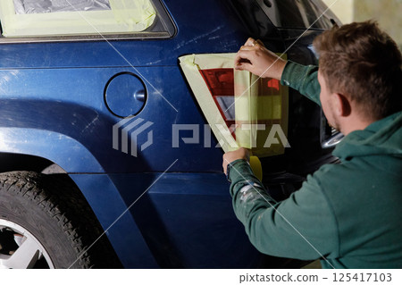 A person meticulously applies masking tape on the rear tail light of a blue SUV in a garage during the evening, preparing for painting and detailing work to enhance the vehicle's appearance. A person meticulously applies masking tape on the rear tail light of a blue SUV in a garage during the evening, preparing for painting and detailing work to enhance the vehicle's appearance. 125417103