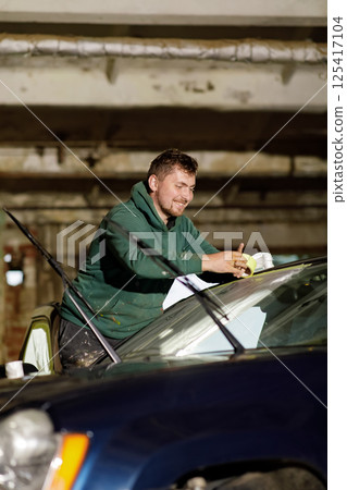 A man works diligently in a garage, cleaning the windshield of a car with a cloth while standing on the vehicle's hood. Natural light filters in from above, highlighting his concentration. 125417104