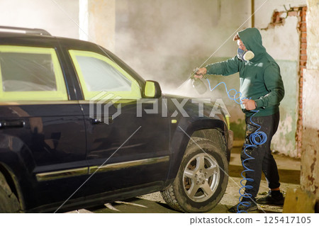 A person wearing a hooded jacket and mask is spray painting a dark-colored car in a garage. The dim light and mist create a focused atmosphere for the detailing work. 125417105