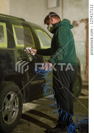 A car painter is carefully applying spray paint to a vehicle's side in a garage setting during the evening. The painter wears protective gear and uses a spray gun while handling the air hose. 125417112