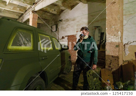 A worker is wearing a mask while carefully painting a large vehicle with camouflage colors in a dim, industrial space illuminated by warm light. Cardboard pieces and tools are nearby. 125417150