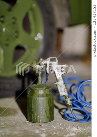 A spray gun attached to a green paint can rests near a green vehicle, with blue air hoses lying on the workshop floor as the light from outside creates a soft ambiance. 125417152