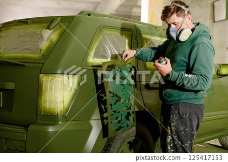 A person is applying camouflage patterns to a green SUV while wearing a mask and protective gear. The setting is a well-lit warehouse with paint supplies nearby. 125417153