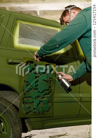 A person is working on a vehicle, using a stencil to add camouflage patterns with spray paint. The setting is a well-lit garage, showcasing a unique customization project for the car. 125417154