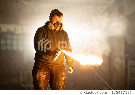 An artist wearing a mask ignites a spray can, creating a vibrant flame in an eerie, abandoned warehouse at dusk. Dust dances in the air, enhancing the dramatic effect of the scene. 125417155