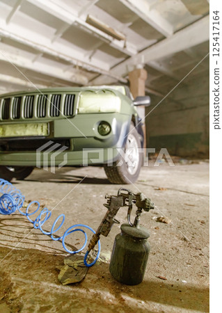 A green vehicle is parked in a garage with faded walls and debris. A spray gun and air hose lay on the ground, suggesting ongoing repair work in a dusty and unkempt environment. 125417164