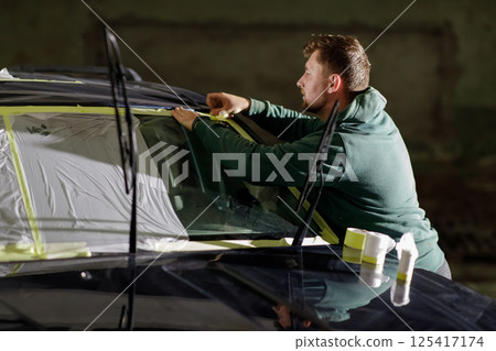 A car detailing professional is focused on taping areas of a vehicle before painting. The workspace is dim, highlighting the detailed preparation involved in automotive care. 125417174