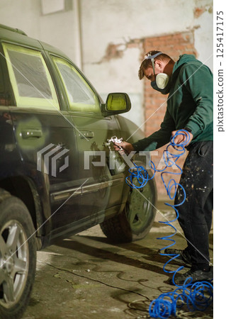 A car painter works diligently in a workshop, using a spray gun to apply paint to a vehicle. The workspace is filled with tools, highlighting the process of automotive refinishing. 125417175