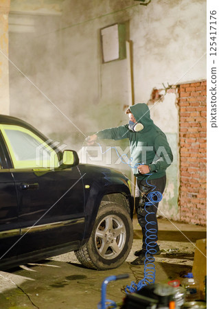 A person wearing a hoodie and safety mask sprays paint on a black vehicle in a dimly lit industrial garage. The atmosphere is filled with mist from the spray paint. 125417176