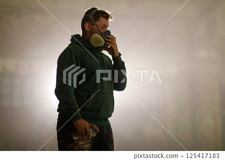 In a studio, an artist stands focused while wearing a mask and holding a spray can. The casual outfit suggests a relaxed environment, highlighting the importance of safety during creative work. 125417183
