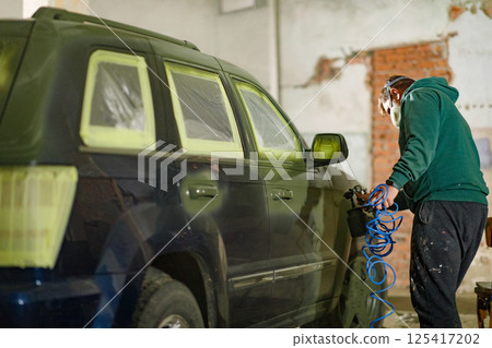 A masked blue vehicle is being painted indoors, with a technician attentively applying finish with a spray gun. The workshop shows exposed brick and prepared surfaces. 125417202